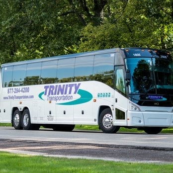 White and blue Trinity Transportation double-decker charter bus parked on a road with trees in the background